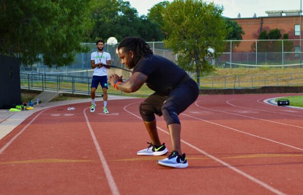 赤い陸上トラックでトレーニングをする人と、その動きを見守る人