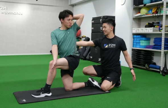 P2M Performanceの室内で、トレーナーのサポートを受けながら片膝立ちで上半身を動かす利用者